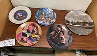 Photo showing six collector plates on wood surface including Dept of the Navy, winter village scene, Tower of London, Norman Rockwell style illustration, Lucy meets Orson Welles, and ship captain portrait.