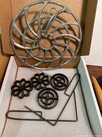 Four metal patty molds and a waffle iron placed in a box with foam padding. The molds include flower and circular shapes, along with metal handle attachments.