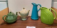 Four items: two ceramic teapots and two ceramic pitchers, various colors and designs, displayed on a wooden surface.