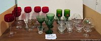 Wide view showing entire collection: red wine glasses, green goblets, small clear glasses with gold trim, decorative decanter and other clear glasses.