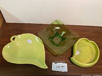 Shows three vintage chartreuse dishes on a wooden surface: a leaf-shaped ceramic dish with handle, a translucent green glass square dish, and a grape-shaped ceramic dish.