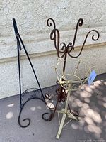 Overview of three wrought iron plant stands against light wall on concrete ground, showing design and color variety.
