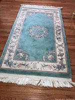Full view of the Chinese area rug showing the central light blue area with floral medallion and cream floral border with fringed ends, laid on hardwood floor