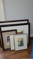 Six framed art pieces leaning against a wall showing diversity of frame styles and artwork sizes.