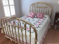 Overall view of the brass full size bed with floral bedding and pink decorative pillows in a bedroom setting with window shutters.