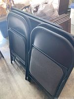 Four black folding chairs with padded seats arranged close together, showing the back and seats, fabric patterned in black and gray.