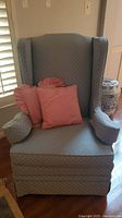 Front view of blue upholstered wingback chair with two pink throw pillows on seat, chair positioned near window with plantation shutters.
