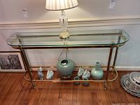 Front view of glass and brass console table with decorative items underneath and a lamp on top (lamp excluded from sale).