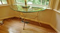 Demi lune shape brass table with clear beveled glass top, empty surface, positioned against a window in a room with hardwood floors.