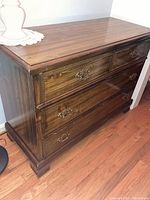 Full front and slight angled view of the wooden dresser showing all three drawers, decorative metal handles, and the glossy wood finish. Drill holes visible on top surface.