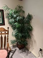 Full view of tall artificial plant near wall and wooden chair, in a woven basket.