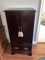 Closed front view of dark wood jewelry cabinet with two panel doors and lower drawer with brass handles.