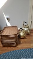 Photo showing stack of 12 rectangular wicker serving trays next to two brass tea kettles on wooden floor