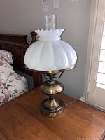 Table lamp with rounded white milk glass shade featuring scalloped edges, brass base, and clear cylindrical chimney on top.