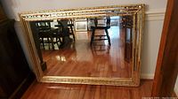 Full view of the gold tone framed beveled mirror resting on the floor, showing its shape and reflection of a room with hardwood floor.