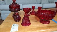 Overall set of ruby red glassware including candy dishes, bell, decanter, and candlesticks on a wooden table with blue tape.