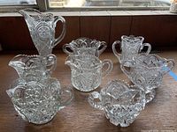 Eight assorted vintage cut glass vessels including sugar bowls, creamers, and a tall pitcher placed on a wooden surface. Detailed starburst and scalloped edge patterns visible.