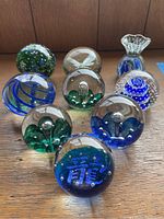 Nine assorted vintage paperweights displayed on wooden surface, showing varied shapes, colors in blue and green shades, includes sphere and flower shaped designs.