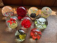 Overview of nine vintage glass paperweights arranged in a group on wooden surface, showing various designs in red and yellow color themes.