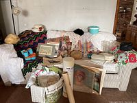 Wide view showing entire sofa area with afghan, doilies, plastic containers, artwork, and various items staged on couch and surrounding area.