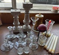 Overview of assorted candle holders and candles displayed on wood surface by window showing wooden pillar candle holders, glass candle holders, and multiple candles.