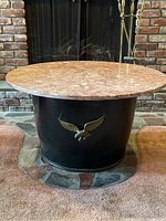 Front view of the metal barrel base table with round pink marble top on carpet in front of fireplace with clear view of 3D metal eagle attached to barrel base and natural marble veining.
