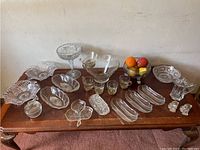 Wide angle view showing entire assortment of vintage clear glass tableware arranged on wooden table beneath plain wall, including chip and dip bowls, candy dishes, pedestal bowls, and drinking glasses.