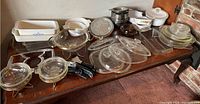 Overview photo of all vintage CorningWare items arranged on a wooden table, showing the variety of glassware, baking dishes, lids, and accessories.