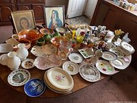 Wide view of the table filled with various vintage dining room items including glassware, plates, framed artwork, and decorative objects.