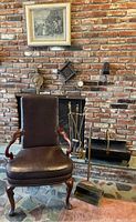 Wide view of fireplace area including leather armchair, metal fireplace tools, log holder, and decorative items on mantel with framed etching above.