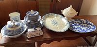 Wide view of all vintage china items on wooden table showing Wedgwood cloche platter, blue willow dishes, butter dish, flow blue platter and Nasco soup tureen.
