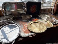 Photo showing assortment of vintage metal serving trays, wire baskets, and hand-painted floral trays arranged on a wooden surface