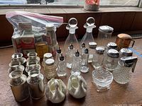 Photo showing overall lot on table near window with various glass, ceramic, metal salt and pepper shakers, oil and vinegar bottles, and a plastic bag of vintage spice cans and packets.