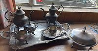 Eight-piece vintage silverplated tea service set displayed on a windowsill, including two teapots, a tray, and other serving pieces, showing tarnish and wear.