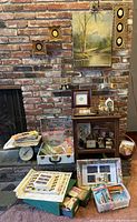 Wide shot showing assortment of vintage framed artwork, stereopticon slides, file cabinet, and postal stamp albums with folders and boxes on a brick fireplace hearth.