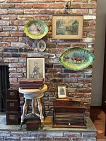 Wide shot of items arranged against a brick wall and fireplace hearth, including piano seat, boxes, framed artwork, and decorative plates.