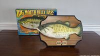 Big Mouth Billy Bass mounted on wooden plaque with original box behind.