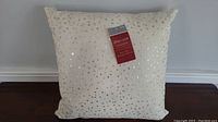 Photo of a square cream-colored pillow with gold and silver sequins scattered on the surface, tag attached in the front center showing brand and dimensions.