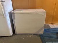 Front view of the white Coldspot chest freezer showing flat top and side handle, located beside a refrigerator in a basement room with bare floor and walls.