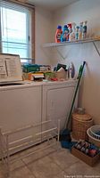 Photo showing metal shoe rack, wicker waste basket with lid, laundry detergent, T-Fal steam iron on dryer and various cleaning supplies on shelf.