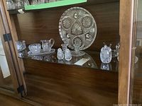 Display of vintage crystal serving tray, salt and pepper shakers, cream and sugar set with tray, candle holders, small bowl inside a wooden cabinet