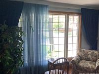 Blue sheer and royal blue pleated panel curtains on window with light coming through, next to floral patterned armchair and wooden chair with glass side table.
