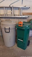 Full view of two large garbage cans, one beige Rubbermaid on wheels and one green can. The beige can has a locking lid and handles.