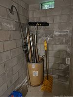 Photo of garden tools and sports equipment stored inside and around a large brown cylindrical storage drum. Shows various metal poles, shovel with yellow handle, and other items in basement corner.