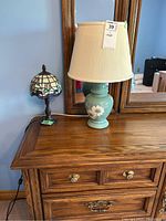 Photo of two lamps on wooden desk with a large mirror behind. Small Tiffany style stained glass lamp with floral design and larger green ceramic lamp with cream shade.