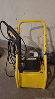 Front view of the Karcher pressure washer showing the black control panel area on yellow frame and attached spray gun with hose coiled on the side.