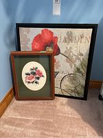 Two framed floral prints shown on floor against blue wall: a larger square black-framed poppy and butterfly print and a smaller wooden-framed pink flower print.
