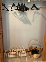 Photo showing black plastic coat hangers hanging from a rod, a large light brown wicker basket with handle on the floor, and a white metal 3 tier shoe rack beside the basket.