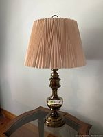 Photo of one brass look table lamp with beige pinched pleat shade on a glass topped wooden table.