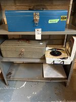 Shelf with blue metal toolbox on top labeled 19x6x6, grey metal toolbox below it which is locked shut, corded drill in case beside grey toolbox.